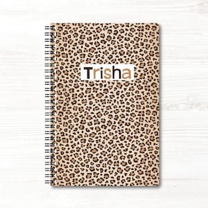 May include: A spiral-bound notebook featuring a tan and brown leopard print pattern across the cover. A white rectangular label is centred on the front with the name Trisha printed in dark brown sans-serif capital and lowercase letters.