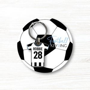 K&ouml;nnte beinhalten: Ein Fu&szlig;ball-Schl&uuml;sselanh&auml;nger mit einem Fu&szlig;ball-Design. Der Schl&uuml;sselanh&auml;nger enth&auml;lt ein Miniatur-Trikot mit dem Namen "Robbie" und der Nummer "28". Die Worte "Football KEYRING" sind hellblau geschrieben.