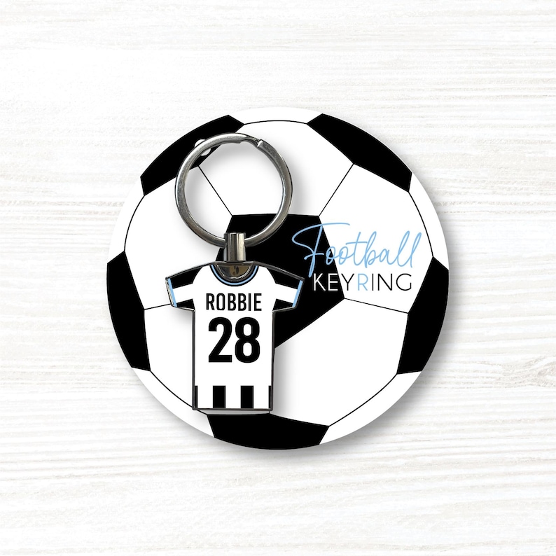 K&ouml;nnte beinhalten: Ein Fu&szlig;ball-Schl&uuml;sselanh&auml;nger mit einem Fu&szlig;ball-Design. Der Schl&uuml;sselanh&auml;nger enth&auml;lt ein Miniatur-Trikot mit dem Namen "Robbie" und der Nummer "28". Die Worte "Football KEYRING" sind hellblau geschrieben.