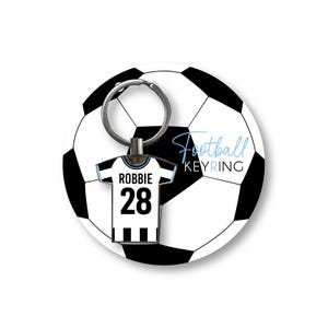 K&ouml;nnte beinhalten: Ein Fu&szlig;ball-Schl&uuml;sselanh&auml;nger mit einem schwarz-wei&szlig;en Fu&szlig;ball-Design. Der Anh&auml;nger hat einen Trikot-f&ouml;rmigen Charm mit dem Namen "Robbie" und der Nummer "28". Der Text "Football KEYRING" ist ebenfalls zu sehen.