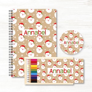 May include: A spiral-bound notebook, a circular coaster, and a wooden box of colored pencils, all featuring a tan background with a repeating pattern of Santa Claus faces, white snowflakes, and the name Annabel printed in red and green letters.