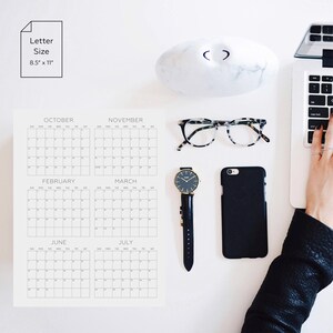 May include: A letter size 8.5" x 11" calendar sheet featuring grids for October, November, February, March, June, and July. Nearby are a white marble paperweight, tortoiseshell glasses, a black wristwatch, a black smartphone, and a laptop.