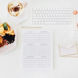 May include: A white desk setup featuring a six-month calendar page for August through January, a white wireless keyboard, a gold pen, a cup of coffee with foam, a plate of sliced peaches and blueberries, and a white envelope with gold trim.