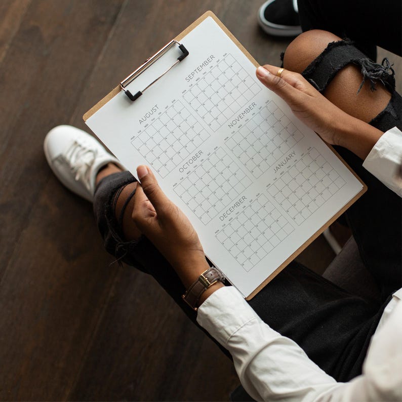 May include: A wooden clipboard holding a white paper sheet printed with a six-month calendar grid. The calendar displays the months of August, September, October, November, December, and January, each organized into a grid of numbered days.
