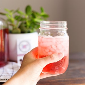 May include: A clear glass mason jar filled with a vibrant red liquid and ice cubes. The jar features a textured glass surface and a threaded rim. In the background, a large glass pitcher containing a similar red beverage sits next to a green plant.