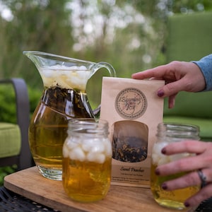 May include: A glass pitcher filled with iced tea and tea leaves, two mason jars filled with iced tea, and a brown paper bag with the text "Blooming with Joy" and "Sweet Peaches" on it. The tea is a loose leaf blend of green tea, peach, and apricot.