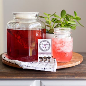 May include: A large square glass pitcher filled with deep red tea, a smaller glass mason jar containing pink iced tea with ice cubes, and a rectangular business card with the text Blooming With Joy and images of tea tins.
