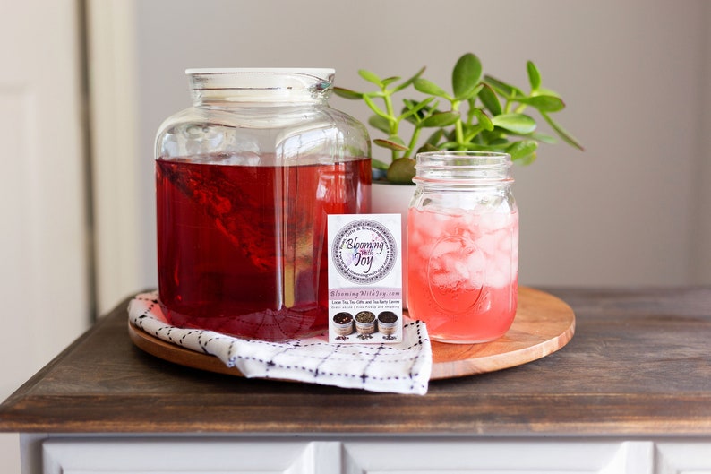 May include: A large square glass pitcher filled with deep red tea, a smaller glass mason jar containing pink iced tea with ice cubes, and a rectangular business card with the text Blooming With Joy and images of tea tins.