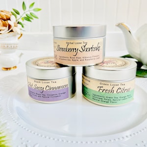 May include: Three silver tins of loose leaf tea are stacked on a white plate. The top tin reads "Strawberry Shortcake," the middle tin says "Hot Sassy Cinnamon," and the bottom tin says "Fresh Citrus." Each tin has a label with the tea type and ingredients.