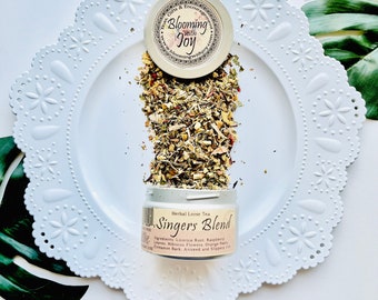 Könnte beinhalten: Ein weißer Keramikteller mit dekorativem Rand, auf dem eine kleine Dose mit loser Kräutertee mit der Aufschrift "Singers Blend" zu sehen ist. Der Tee rieselt aus der Dose und zeigt eine Mischung aus getrockneten Kräutern und Blüten. Die Dose hat ein Etikett mit dem Text "Herbal Loose Tea Singers Blend" und einer Zutatenliste: Süßholzwurzel, Himbeerblätter, Wildblumen, Orangenschalen, Zimtrinde, Anis und Ulmenrinde.