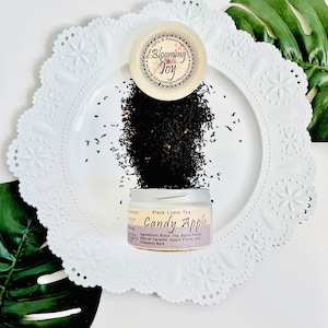 May include: A white ceramic plate with a scalloped edge, a small tin of black loose leaf tea labeled "Candy Apple" and the text "Black Loose Tea" and a list of ingredients. The tin is open and the tea is spilling out onto the plate.