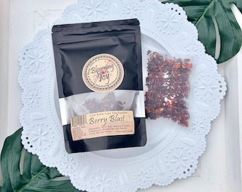 May include: A black pouch of Blooming with Joy Berry Blast cold brew iced tea with a circular logo and a label listing ingredients like rose hips, hibiscus flowers, cranberries, and blueberries. A small clear bag of dried tea leaves sits beside it.