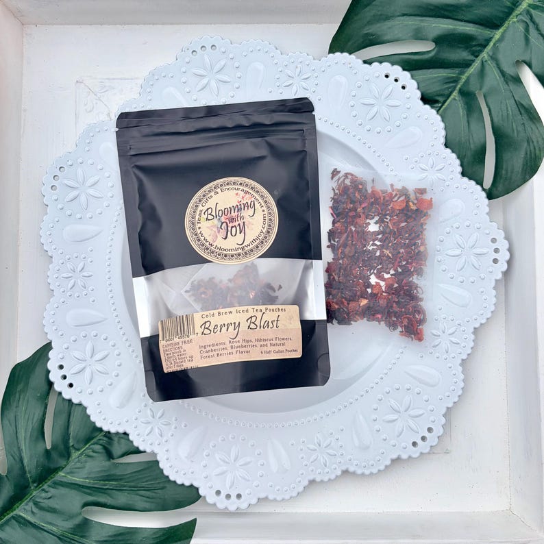 May include: A black pouch of Blooming with Joy Berry Blast cold brew iced tea with a circular logo and a label listing ingredients like rose hips, hibiscus flowers, cranberries, and blueberries. A small clear bag of dried tea leaves sits beside it.