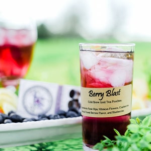 May include: A clear glass filled with dark red iced tea and ice cubes, featuring a tan label that reads Berry Blast Cold Brew Iced Tea Pouches, with ingredients listed as Rose Hips, Hibiscus Flowers, Cranberries, Natural Forest Berries Flavor, and Blueberries.
