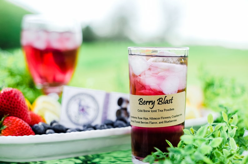 May include: A clear glass filled with dark red iced tea and ice cubes, featuring a tan label that reads Berry Blast Cold Brew Iced Tea Pouches, with ingredients listed as Rose Hips, Hibiscus Flowers, Cranberries, Natural Forest Berries Flavor, and Blueberries.