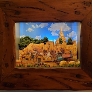 May include: A framed diorama depicting a desert landscape with a train. The scene includes a mountain range, green trees, and a blue sky with clouds. The frame is made of rustic, dark brown wood.