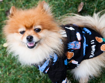 May include: A small, fluffy, orange and white dog wearing a black and blue patterned shirt with the text "HAWAII" on it. The dog is standing on green grass.