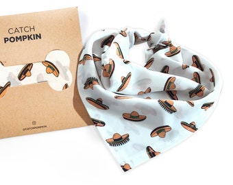 May include: A light blue bandana with a pattern of brown sombreros on a white background. The bandana is folded and tied in a knot. The bandana is packaged in a brown paper envelope with a bone-shaped cutout and the text "CATCH POMPKIN" printed on it.