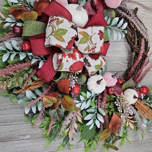 Op de afbeelding: Een decoratieve krans met een basis van wijnranken, versierd met groen blad, kleine witte pompoenen, rode bessen en gedroogd gras. Een grote, meerlaagse strik van bordeauxrood lint en lint met een patroon van herfstbladeren siert de bovenkant.