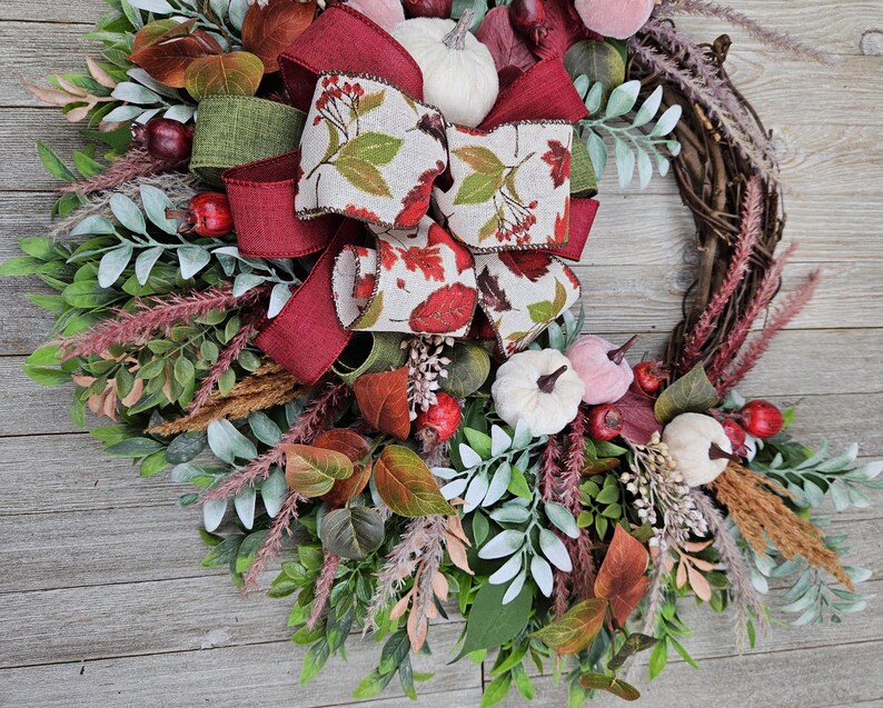 Op de afbeelding: Een decoratieve krans met een basis van wijnranken, versierd met groen blad, kleine witte pompoenen, rode bessen en gedroogd gras. Een grote, meerlaagse strik van bordeauxrood lint en lint met een patroon van herfstbladeren siert de bovenkant.