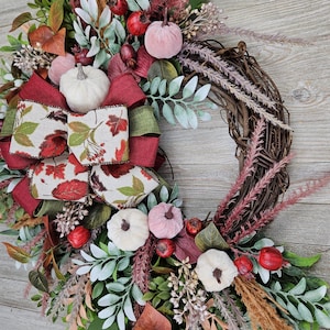 Op de afbeelding: Een decoratieve krans met een basis van bruine takken, versierd met kleine witte en roze pompoenen, rode bessen, groen blad en gedroogd gras. Een grote, meerlaagse strik van rode stof en cr&egrave;mekleurig lint met een herfstbladpatroon.