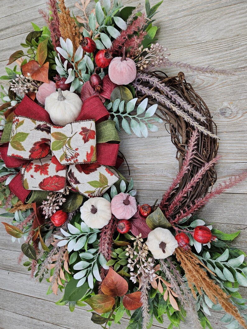 Op de afbeelding: Een decoratieve krans met een basis van bruine takken, versierd met kleine witte en roze pompoenen, rode bessen, groen blad en gedroogd gras. Een grote, meerlaagse strik van rode stof en cr&egrave;mekleurig lint met een herfstbladpatroon.