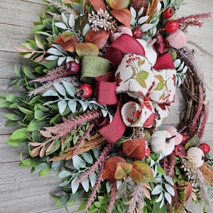 Op de afbeelding: Een decoratieve krans met een basis van druivenranken, versierd met groen blad, gedroogd gras en kleine witte pompoenen. Het arrangement bevat rode bessen en een meerlaagse strik van bordeauxrood lint en cr&egrave;mekleurig lint met een bladprint.