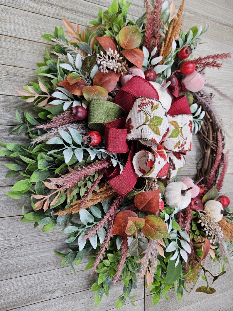 Op de afbeelding: Een decoratieve krans met een basis van druivenranken, versierd met groen blad, gedroogd gras en kleine witte pompoenen. Het arrangement bevat rode bessen en een meerlaagse strik van bordeauxrood lint en cr&egrave;mekleurig lint met een bladprint.
