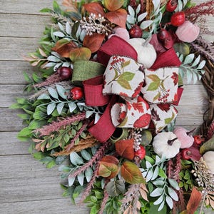 Op de afbeelding: Een decoratieve krans met een mix van groen en saliekleurig blad, gedroogde grassen en kleine pompoenen in wit en gedempt roze. Het arrangement is geaccentueerd met donkerrode bessen en een meerlaagse strik van bordeauxrode en bedrukte stof.