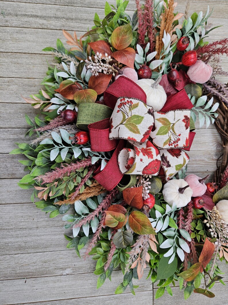 Op de afbeelding: Een decoratieve krans met een mix van groen en saliekleurig blad, gedroogde grassen en kleine pompoenen in wit en gedempt roze. Het arrangement is geaccentueerd met donkerrode bessen en een meerlaagse strik van bordeauxrode en bedrukte stof.