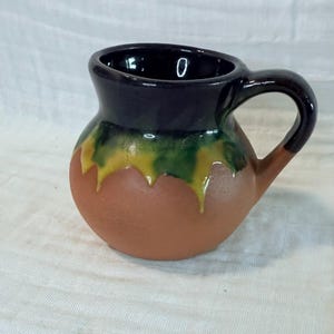 May include: A handmade, round, ceramic mug with a handle. The mug features a dark blue rim, a brown body, and a green and yellow drip design. The handle is dark blue and brown. The mug is likely used for drinking beverages.
