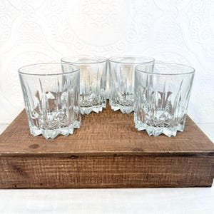 May include: Four clear, faceted drinking glasses with a starburst design on the base. The glasses are arranged on a rustic, brown wooden tray. The glasses have a classic, vintage aesthetic.