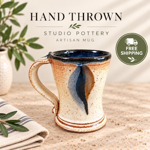 May include: A hand-thrown ceramic mug with a speckled cream glaze, featuring a prominent blue and brown vertical accent on the side, a dark blue interior rim, and a sturdy handle. The text reads HAND THROWN STUDIO POTTERY ARTISAN MUG.