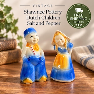 May include: A pair of vintage Shawnee Pottery salt and pepper shakers shaped like a boy and a girl in Dutch-style clothing. The boy wears a blue cap, orange scarf, and blue trousers. The girl wears a tan bonnet, blue bodice, and orange apron.