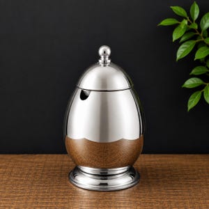 May include: A stainless steel egg-shaped ashtray with a wooden base. The ashtray has a lid with a small ball on top and a small opening for cigarettes. The ashtray sits on a round base.