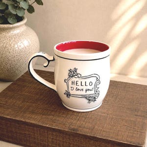 May include: A white ceramic mug with a bright red interior and a black handle. The exterior features a black hand-drawn style frame containing the text "HELLO I love you!" with small floral accents on the corners of the decorative border.