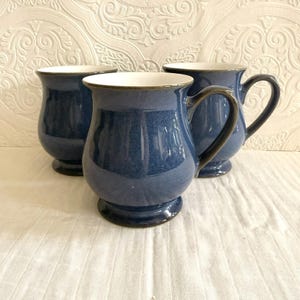 May include: Three blue ceramic mugs with a dark handle and a white interior. The mugs have a rounded shape and sit on a small base. The mugs are arranged on a white surface.