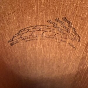 May include: Close-up of a wooden surface with a black graphic. The graphic features the text "CIGAR TRAVELER" in a curved design, with stylized smoke rising above. The background is a warm, brown tone.