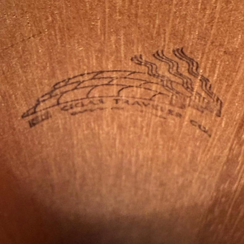 May include: Close-up of a wooden surface with a black graphic. The graphic features the text "CIGAR TRAVELER" in a curved design, with stylized smoke rising above. The background is a warm, brown tone.
