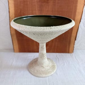May include: A textured, off-white martini glass with a dark green interior. The glass has a wide, conical bowl and a slender stem, resting on a circular base. The surface has a rough, speckled appearance, giving it a unique, handcrafted look.