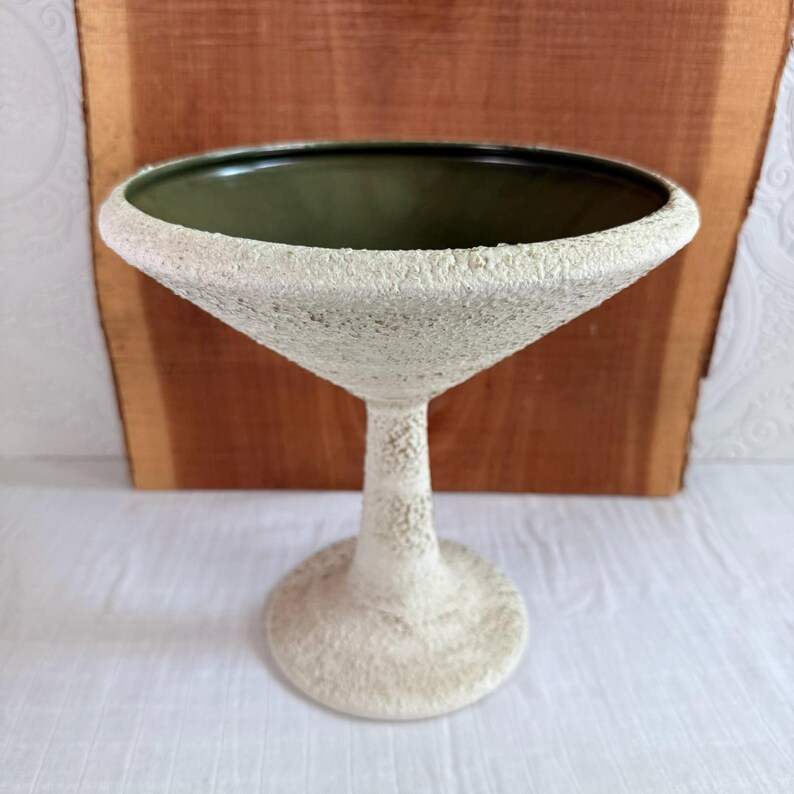 May include: A textured, off-white martini glass with a dark green interior. The glass has a wide, conical bowl and a slender stem, resting on a circular base. The surface has a rough, speckled appearance, giving it a unique, handcrafted look.