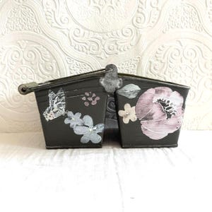 May include: A dark gray metal planter with a handle and two compartments. The planter is decorated with a butterfly, flowers, and a large pink floral design. The planter is designed for decorative use.
