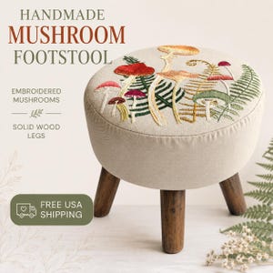 May include: A round footstool with a beige fabric top featuring colorful embroidered mushrooms and green fern leaves. The stool is supported by three dark brown solid wood legs. Text on the image reads HANDMADE MUSHROOM FOOTSTOOL, EMBROIDERED MUSHROOMS, SOLID WOOD LEGS, and FREE USA SHIPPING.