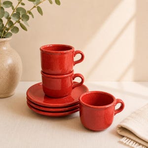 May include: A set of three red ceramic cups and two matching saucers. The cups feature a speckled finish and a rounded handle. Two cups are stacked vertically on top of the saucers, while the third cup sits independently to the right side.