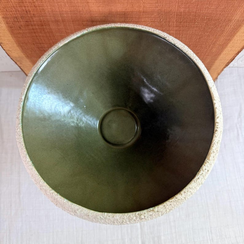 May include: Overhead view of a ceramic bowl with a dark green interior and a textured, off-white rim. The bowl has a circular indentation in the center. The bowl is likely handmade and has a rustic aesthetic.