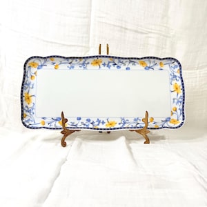 May include: Rectangular white ceramic serving platter with a scalloped edge. The platter is decorated with a blue border and floral pattern in yellow and blue. The platter is displayed on a decorative gold-colored stand.