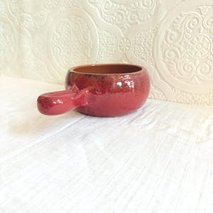 May include: A red ceramic soup bowl with a handle. The bowl has a glossy finish and a textured surface. The handle is attached to the side of the bowl. The interior of the bowl is a darker brown color.