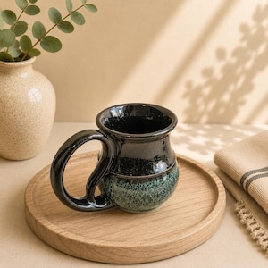 May include: A ceramic mug with a glossy black upper section and a textured, mottled teal and black lower section. The mug features a large, curved handle and a wide rim, showcasing a distinct two-tone glaze finish with a smooth, reflective surface.
