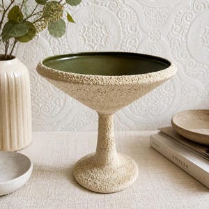 May include: A decorative, textured, off-white ceramic bowl with a wide, flared rim and a dark green interior. The bowl sits atop a pedestal base. The background includes a white vase with greenery and a book.