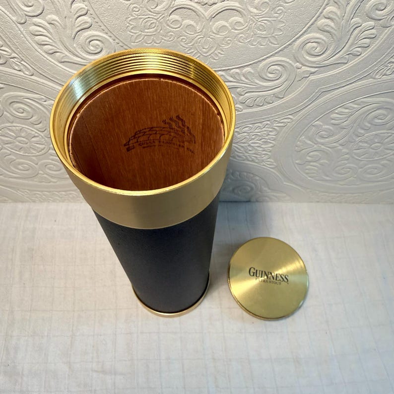 May include: A cylindrical Guinness branded container with a brass-colored rim and lid. The container is black with a wooden interior. The lid is brass-colored and has the Guinness logo.